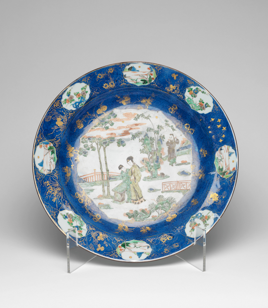 An image of Charger/dish. Unknown production, China. The central medallion has two lovers in a garden with a servant boy looking on from behind ornamental rockwork encircled on the rim with vignettes enclosing flowers and lakeside scenes, and gilding. The shallow rounded sides with broad everted rim resting on a low tapered foot. The interior is reserved against the deep mottled blue ground with a central medallion enclosing a scene of two lovers in a garden picked out in famille verte enamels, the pair are standing beside a stone bench beneath a maple tree with a servant boy looking on from behind a tall outcrop of ornamental rockwork; the medallion is encircled by eight flowering sprays in gilding and the rim has a gilt border of quatrefoil floral vignettes interspersed with famille verte vignettes alternately showing flowers growing behind pierced blue rockwork and lakeside scenes. The underside with three iron-red flowering sprays. The base has two concentric circles in underglaze blue enclosing the mark. Hard-paste porcelain with a underglaze powder-blue ground, famille verte enamels, and gilding, height 6.3 cm, diameter 40.2 cm. Qing Dynasty (1644-1912). Kangxi Period (1662-1722).