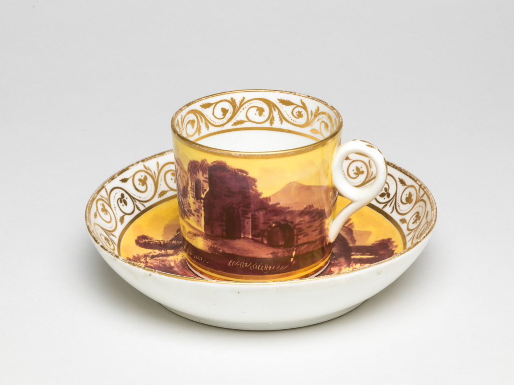 An image of Coffee can and saucer/cup and saucer. Minton factory, Staffordshire, Stoke-on-Trent. The can is cylindrical with a circular ring handle. The saucer has deep curved sides and stands on a footrim. The cup is decorated externally with a yellow ground on which is painted a dark maroon landscape with a ruin on the right, and a low farm house and a tree on the left with mountains in the distance. There is a gold band below and another on the rim. Inside at the top there is a border of gold foliated scrolls. The cup is decorated en suite with ruins in a landscape with trees and distant mountains radiating from two gold concentric circles in the middle. Below the the rim there is a border of foliated scrolls between gold horizontal bands. Pattern no. 604. Bone china (?) decorated with a bright yellow ground, painting in dark maroon enamel, and gilding, height, cup, 6 cm, diameter, cup, 6.8 cm, width, cup, 8.3 cm, height, saucer, 3.2 cm, diameter, saucer, 13.8 cm, circa 1800. Production Note: The decoration imitates French late 18th porcelain, decorated with yellow grounds and monochrome purple or dark puce landscapes. On Minton, the painting is often executed rather roughly.