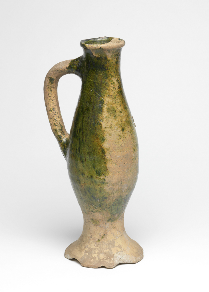 An image of Baluster jug. Unknown potter, England, Surrey, Farnham. Slender oviform body with slightly expanding neck. Loop handle and wide spreading base. Pale buff earthenware, thrown, and partially covered with brown-flecked green lead glaze. Height, whole, 32.3 cm, diameter, foot, 11.9 cm, circa 1275-1325. Medieval.