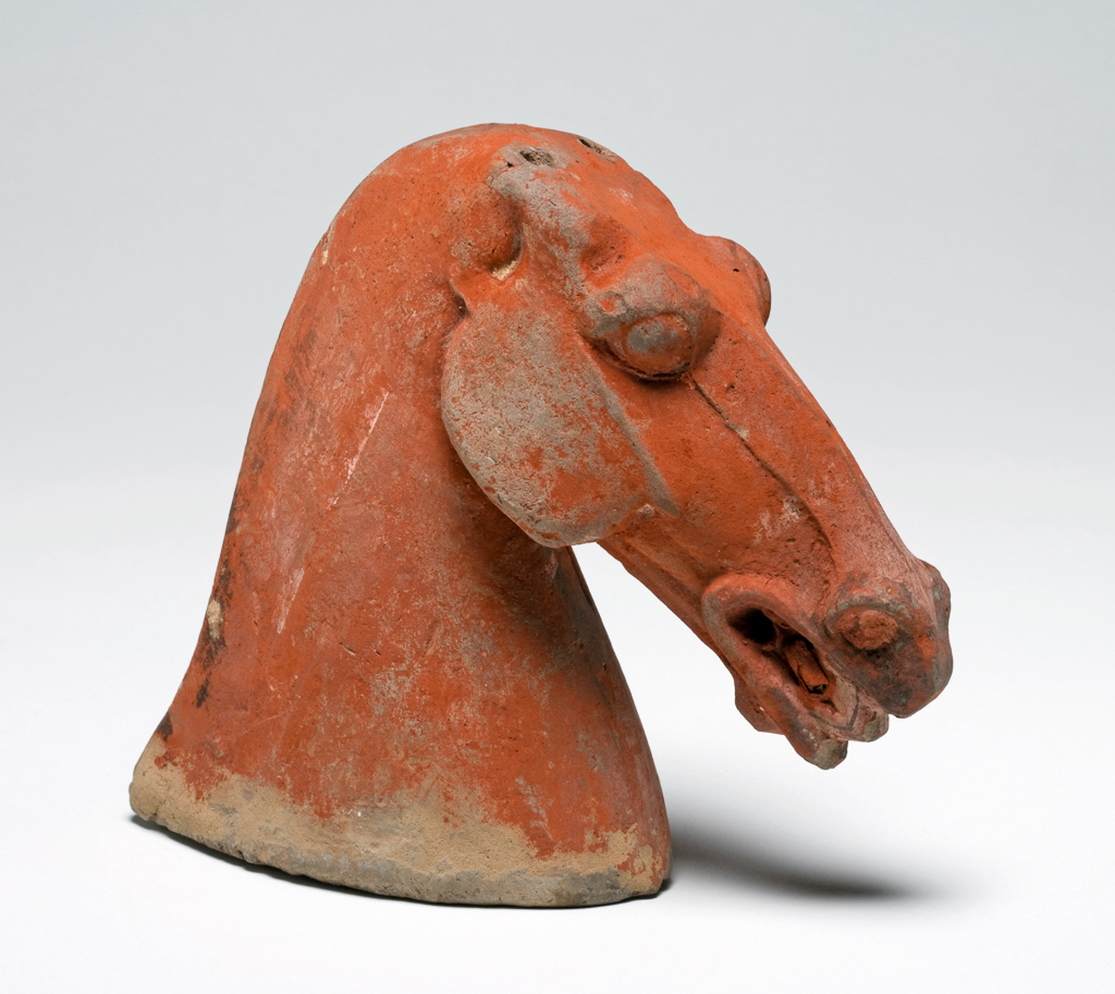 An image of Figure. Horse’s head. Unknown maker, China. Grey earthenware horse head made from two moulds, then joined and seamed. A slip covers the biscuit, and then the figure was painted red with black and white trim. There are thin lines designating teeth. While there are peg holes for ears, the ears themselves are missing. Height 14.6 cm. Hei Dynasty 386-535. Chinese.