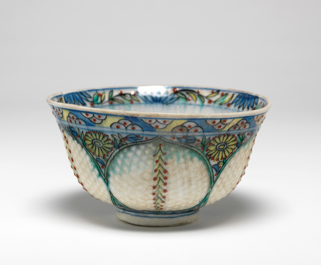 An image of Fritware (stonepaste) bowl. Interior: around the rim a frieze of blue petals and stylised stems with green and yellow leaves interspersed with red dots. Painted in the roundel and enclosed within two blue concentric lines, a polychrome floral spray framed by two green ‘saz’ style leaves. Exterior: on the rim a repeating frieze of three black semi-circles with red central dot is reserved in blue and yellow. On the body inter-locking palmette medallions outlined in green and black contain incised lozenges and a dotted pine stem in yellow, black and red. Glaze covers the exterior surface including the underside of the foot ring. Fritware, wheel made with incised decoration and painted in blue, red, yellow, black and green under a clear, slightly crazed glaze, height, whole, 7.1 cm, width, whole, 12.3 cm, diameter, rim, 12.3 cm, diameter, base, 4.6 cm, weight, whole, 207 g, circa 1700-1750. Ottoman. Islamic pottery. Anatolia, Kutahiya, Kütahya.