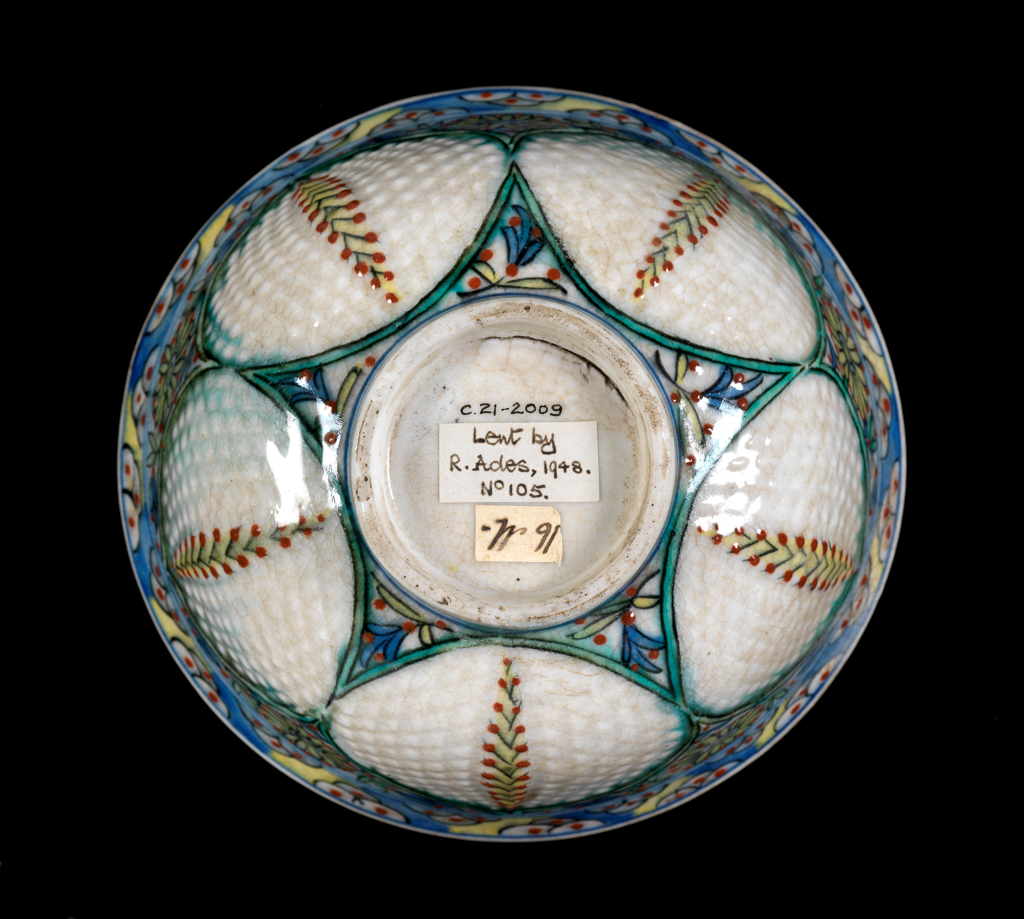 An image of Fritware (stonepaste) bowl. Interior: around the rim a frieze of blue petals and stylised stems with green and yellow leaves interspersed with red dots. Painted in the roundel and enclosed within two blue concentric lines, a polychrome floral spray framed by two green ‘saz’ style leaves. Exterior: on the rim a repeating frieze of three black semi-circles with red central dot is reserved in blue and yellow. On the body inter-locking palmette medallions outlined in green and black contain incised lozenges and a dotted pine stem in yellow, black and red. Glaze covers the exterior surface including the underside of the foot ring. Fritware, wheel made with incised decoration and painted in blue, red, yellow, black and green under a clear, slightly crazed glaze, height, whole, 7.1 cm, width, whole, 12.3 cm, diameter, rim, 12.3 cm, diameter, base, 4.6 cm, weight, whole, 207 g, circa 1700-1750. Ottoman. Islamic pottery. Anatolia, Kutahiya, Kütahya.