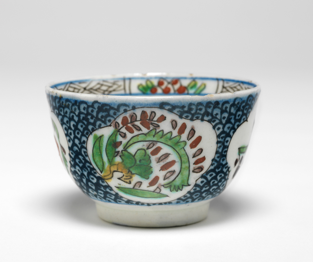 An image of Fritware (stonepaste) cup. Interior: around the rim a frieze alternating between panels of black cross-hatch design and black semi-circles with red dots and green leaves. In the roundel a flower is painted in red and green. Exterior: on the body lobbed medallions containing stylised floral or vegetal designs are reserved against a blue ground covered with a black dotted fish scale pattern. Fritware wheel made and finely potted painted in blue, yellow, red, green, purple and black under a clear glaze, height, whole, 4.7 cm, width, whole, 7.4 cm, diameter, rim, 7.4 cm, diameter, base, 3.8 cm, weight, whole, 50 g, circa 1700-1750. Ottoman. Islamic pottery. Anatolia, Kutahiya, Kütahya.