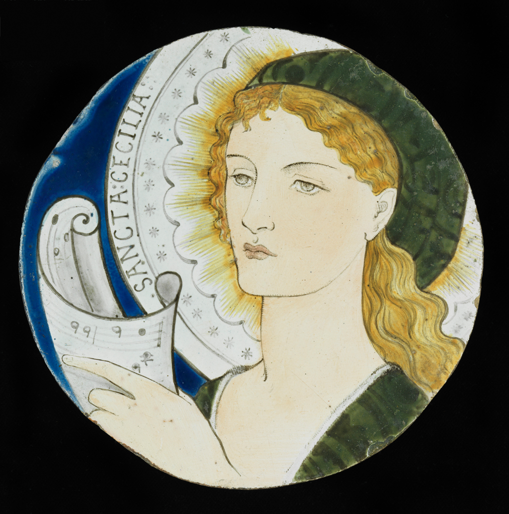 An image of Art Pottery. Tile. Sancta Cecilia. Burne-Jones, Edward (British, 1833-1898). Morris, Marshall, Faulkner & Co. Faulkner, Kate, painter (British, 1842-1898). A head and shoulders painted portrait of a woman, drawn in black, set on a deep blue ground. Her long, wavy, blonde hair is tucked behind her ear and she wears a green cap and gown. She holds a scroll of music in her right hand. Around her head is an elaborate halo, inscribed 'SANCTA CECILIA'. The glaze is dull, the sides of the tile are unglazed and the back is flat and rough. Cut down 6 inch tile. Buff earthenware, slip-coated, glazed and painted in blue, yellow, sage green, flesh-pink and black enamel colours, diameter 15.2 cm, circa 1865. Arts and Crafts. Notes: Saint Cecilia is the patron saint of musicians and Church music.