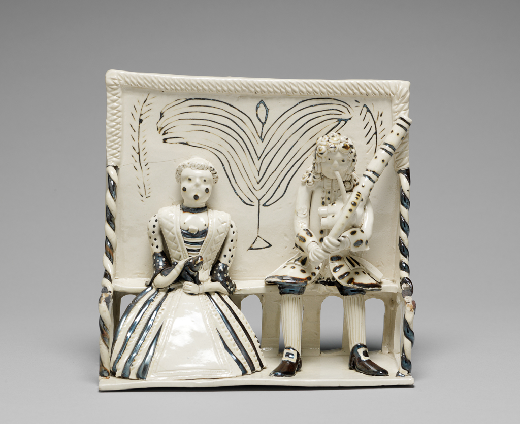 An image of Stoneware / Figure Group. Pew Group. Unknown. Staffordshire factory. White salt-glazed stoneware with details in brown, height, whole, 16.5 cm, width, whole, 16.5 cm, circa 1740-1750. George II. Pew group of a woman holding a dog, and a man playing a musical instrument, both seated on a high-backed settle. Hand-modelled.