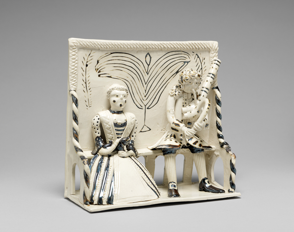 An image of Stoneware / Figure Group. Pew Group. Unknown. Staffordshire factory. White salt-glazed stoneware with details in brown, height, whole, 16.5 cm, width, whole, 16.5 cm, circa 1740-1750. George II. Pew group of a woman holding a dog, and a man playing a musical instrument, both seated on a high-backed settle. Hand-modelled.