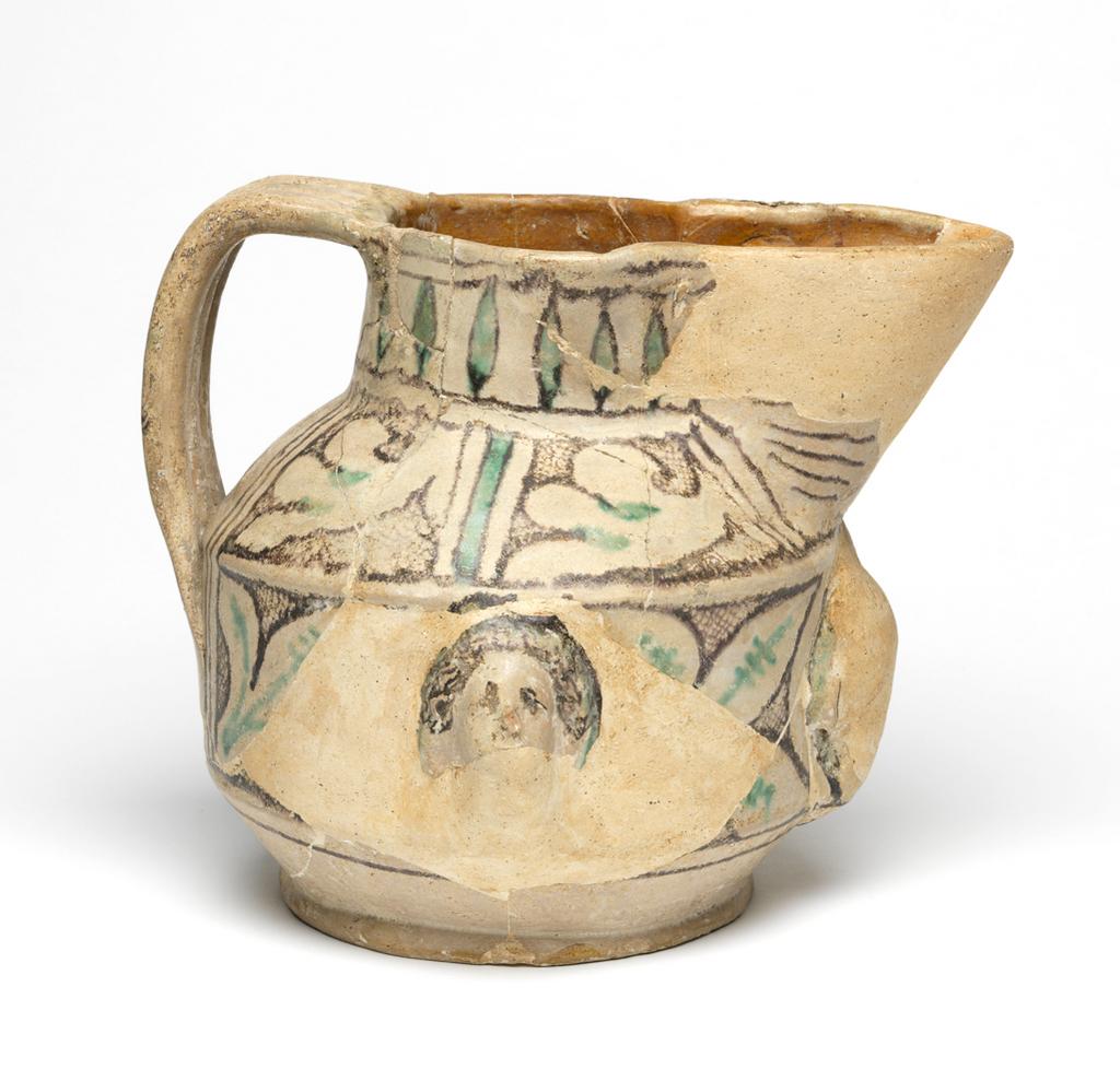 An image of Maiolica arcaica. Jug. Unidentified Orvieto pottery, Italy, Umbria. Shape 10. Thickly potted bulbous body with vertical zone round the widest part, cylindrical neck with large pelican-beak spout, and wide strap handle composed of three rolls of clay pressed together and flattened. The jug originally had relief heads on the front and sides, but only one of the latter remains. The vertical area is decorated with leaves, with two manganese bands below, and each side of the shoulder, with two panels filled with leaves and cross-hatching. On the neck, there are lanceolate leaves between two pairs of manganese bands; on the spout, manganese horizontal lines; and on the handle, three groups of manganese stripes alternating with two green, with three green below. Earthenware: the interior is lead-glazed yellowish-brown, the exterior is tin-glazed pale greyish-white; painted in manganese and copper-green. Height, whole, 20.4 cm, diameter, base, 14.1 cm, width, handle to spout, 24.5 cm, circa 1300-1400. Late Medieval.