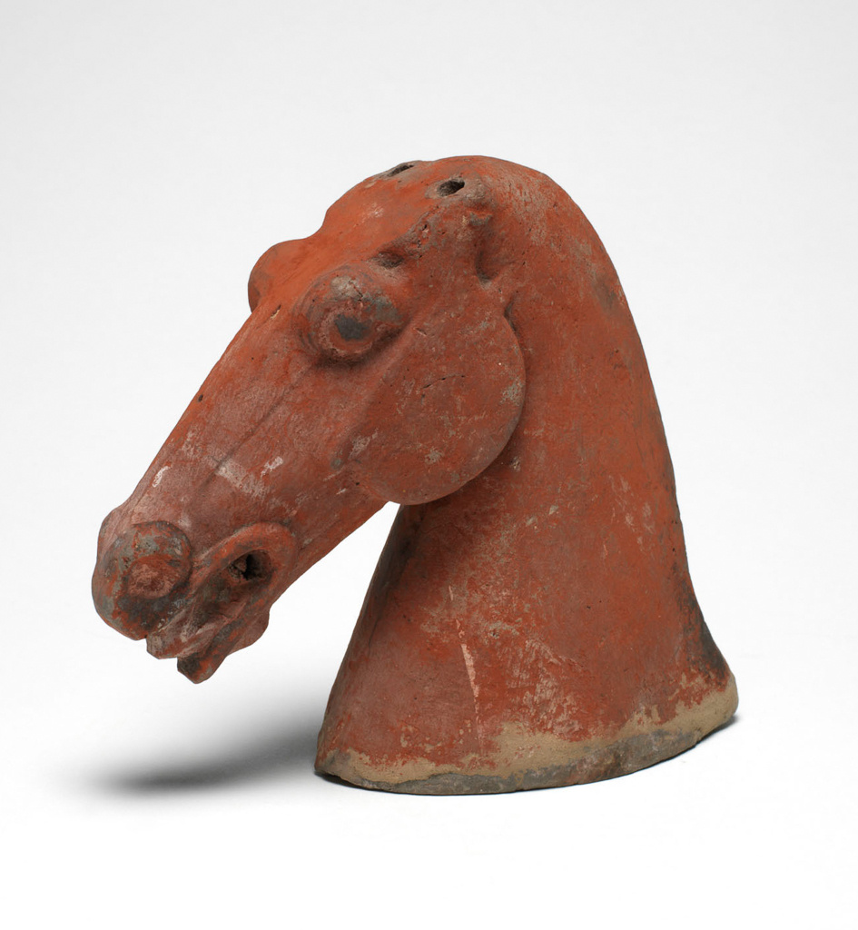 An image of Figure. Horse’s head. Unknown maker, China. Grey earthenware horse head made from two moulds, then joined and seamed. A slip covers the biscuit, and then the figure was painted red with black and white trim. There are thin lines designating teeth. While there are peg holes for ears, the ears themselves are missing. Height 14.6 cm. Hei Dynasty 386-535. Chinese.