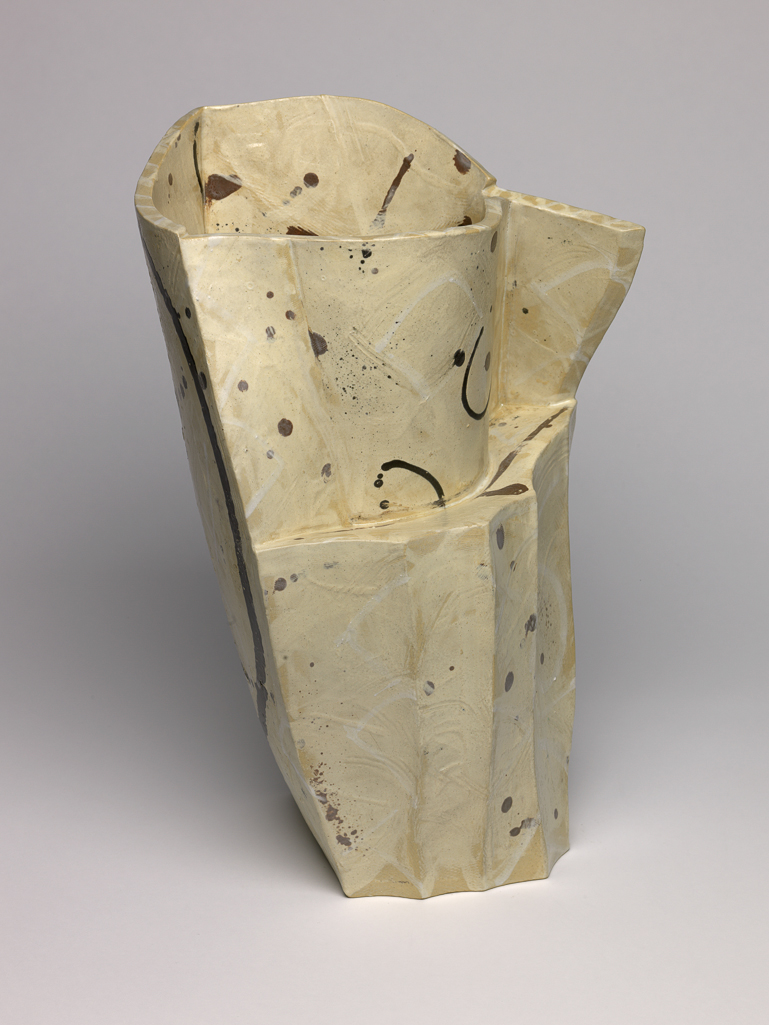 An image of Studio Ceramics. White Pot, Brown Dots. Britton, Alison (British, b. 1948). Tall slab-built vessel, rising from a flat base to a wider opening at the top, both near-triangular in shape. Two sides are each built from a single piece of clay, the third formed from several pieces in a mix of angular and curving shapes. The surface is coated and painted with slips, with gestural marks and dots in white and brown over varied buffs. The decoration, mostly applied prior to fabrication, displays the physical marks of application. The underside is flat and unglazed. Earthenware, slab-built and painted with slips and matt glazed. Height 41.5 mm, width 26.5 mm, depth 23 mm, 1986.