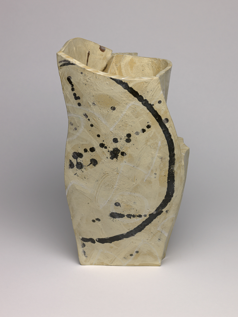 An image of Studio Ceramics. White Pot, Brown Dots. Britton, Alison (British, b. 1948). Tall slab-built vessel, rising from a flat base to a wider opening at the top, both near-triangular in shape. Two sides are each built from a single piece of clay, the third formed from several pieces in a mix of angular and curving shapes. The surface is coated and painted with slips, with gestural marks and dots in white and brown over varied buffs. The decoration, mostly applied prior to fabrication, displays the physical marks of application. The underside is flat and unglazed. Earthenware, slab-built and painted with slips and matt glazed. Height 41.5 mm, width 26.5 mm, depth 23 mm, 1986.