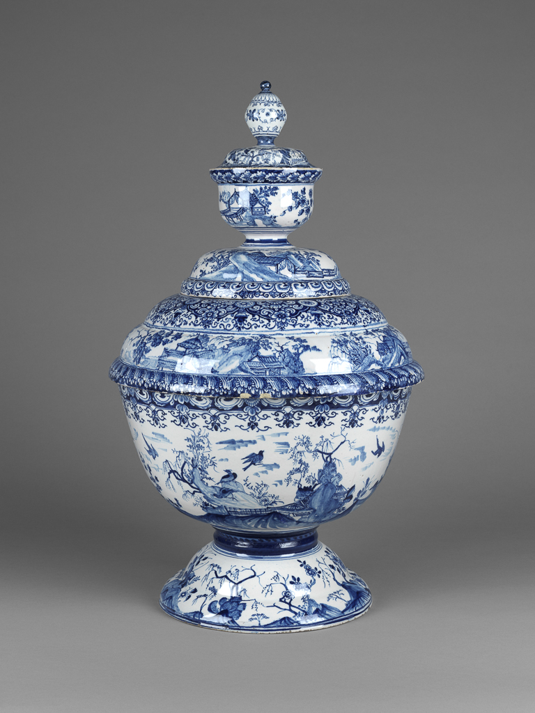 An image of English Delftware. Punch bowl with three-part cover. Liverpool Factory. The bowl itself rests on a high domed foot and is painted round the outside with a Chinese landscape, with pavilions on rocks, trees and flying birds, below a border of formal foliage; a similar landscape on the foot. Inside, the arms of Liverpool with the motto 'DEUS.NOBIS.HAEC.OTIA.FECIT'; symmetrical scroll ornaments round the side. The lower part of the lid is domed and painted with similar Chinese landscapes below a panelled border; it has in the middle of it a deep depression forming a small bowl which is covered by the second lid; this supports a third small receptacle, also covered by means of the uppermost lid which ends in a bulbous knob (whole lid is a replacement). Both upper lids are painted with Chinese landscapes; the lower one of them is inscribed inside: 'THOMAS BOOTLE ESQUIRE MEMBER OF PARLIAMENT FOR LIVERPOOLE 1724'. Earthenware, thrown in parts, tin-glazed and painted in cobalt blue, height, whole, 59 cm, diameter, bowl, 35.5 cm, dated 1724. Oriental Style.