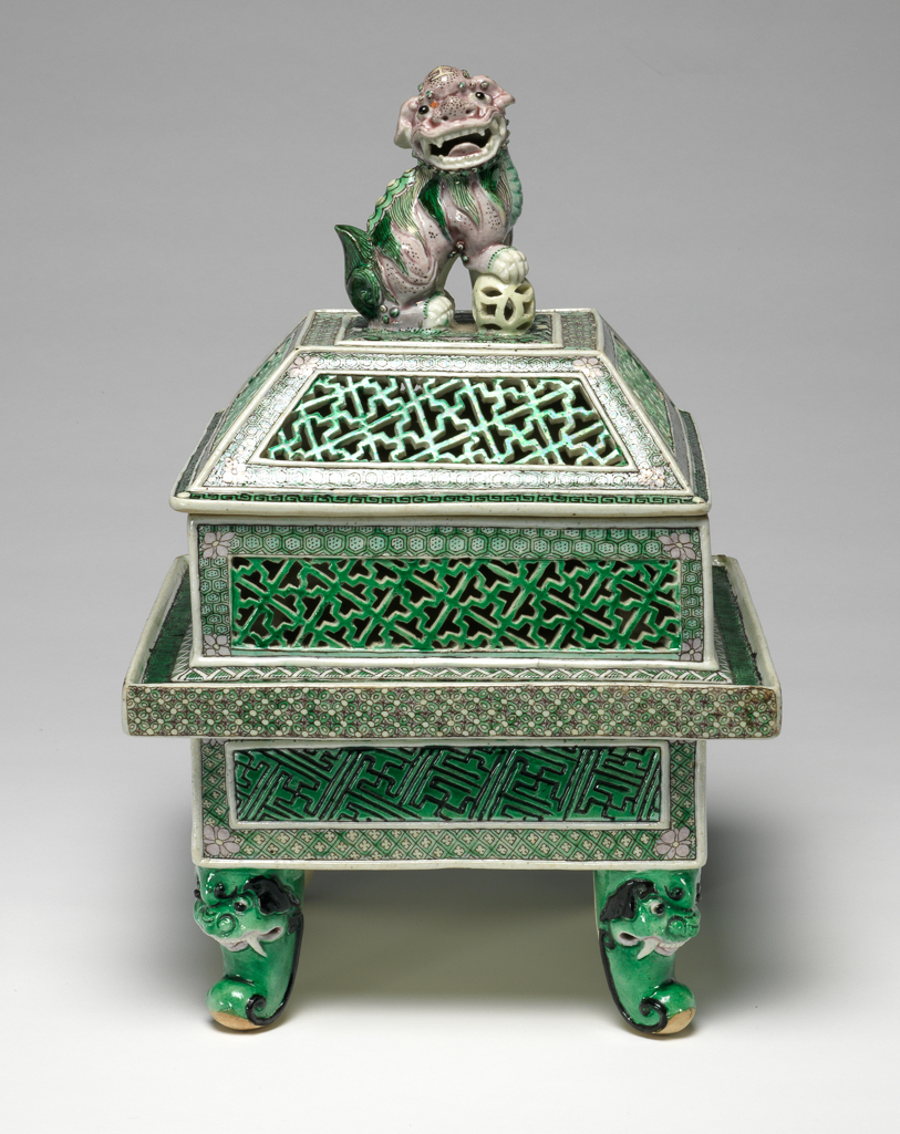 An image of Perfume burner and cover/cassolette. Famille verte, biscuit. The two-storied body is of square section with upright sides supported on four green scroll legs issuing from ferocious animal masks. The lower part of the body has raised rectanglar panels incised with a wan-diaper pattern picked out in black on a green ground, reserved against an intricate green and yellow trellis diaper, surmounted by a galleried flange with a florette diaper in green, yellow and aubergine round the edge and a green-ground spiral scroll on the top. The upper part of the body is intricately pierced with green wan-diaper panels reserved against pale green cell-diaper. There is a matching pierced wan-diaper and cell-diaper pattern on the sloping sides of the domed cover which has a flat top, centred with a large pale aubergine Buddhist lion knop, the animal's right paw resting on a pale yellow brocade ball. The interior and the base are unglazed. Hard-paste porcelain, pierced and applied, and painted on the biscuit in an enamel palette of black, yellow, aubergine and two tones of green, height, whole, 29.5 cm, diameter, whole, 19.6 cm, circa 1662-circa 1722. Late 19th century. Qing Dynasty (1644-1912). Chinese.