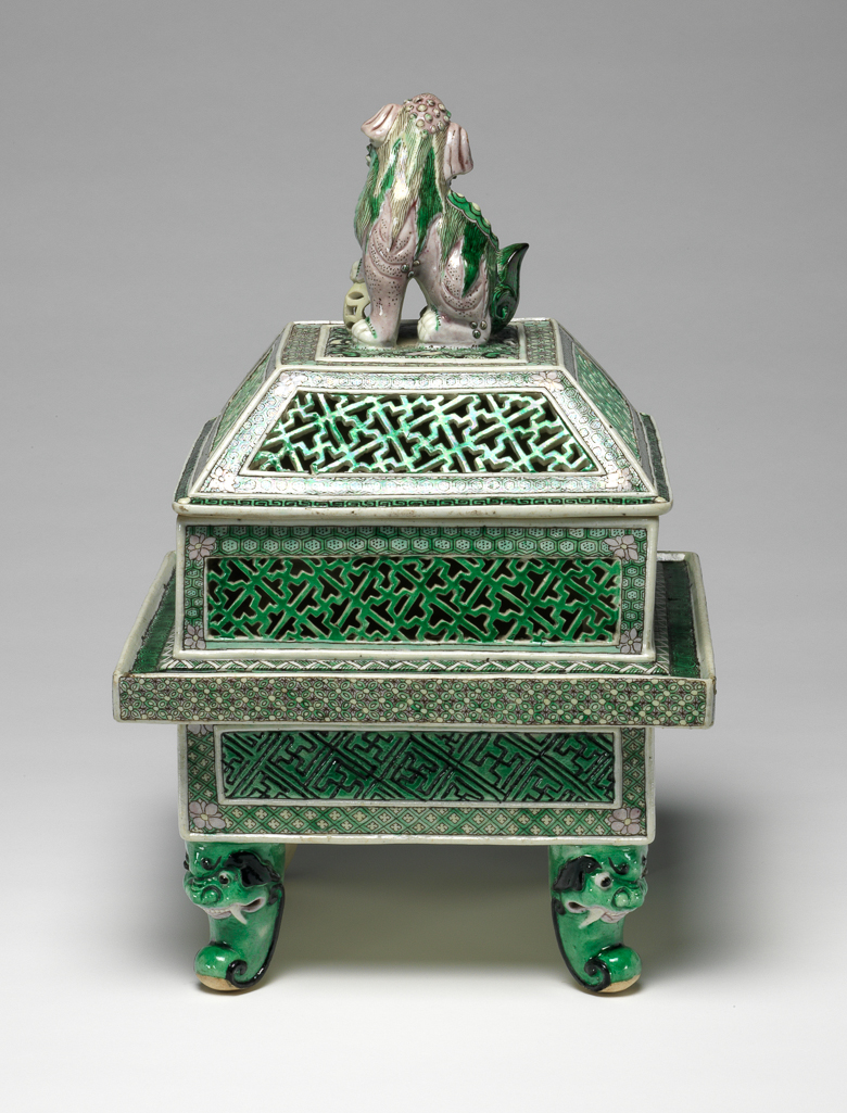 An image of Perfume burner and cover/cassolette. Famille verte, biscuit. The two-storied body is of square section with upright sides supported on four green scroll legs issuing from ferocious animal masks. The lower part of the body has raised rectanglar panels incised with a wan-diaper pattern picked out in black on a green ground, reserved against an intricate green and yellow trellis diaper, surmounted by a galleried flange with a florette diaper in green, yellow and aubergine round the edge and a green-ground spiral scroll on the top. The upper part of the body is intricately pierced with green wan-diaper panels reserved against pale green cell-diaper. There is a matching pierced wan-diaper and cell-diaper pattern on the sloping sides of the domed cover which has a flat top, centred with a large pale aubergine Buddhist lion knop, the animal's right paw resting on a pale yellow brocade ball. The interior and the base are unglazed. Hard-paste porcelain, pierced and applied, and painted on the biscuit in an enamel palette of black, yellow, aubergine and two tones of green, height, whole, 29.5 cm, diameter, whole, 19.6 cm, circa 1662-circa 1722. Late 19th century. Qing Dynasty (1644-1912). Chinese.