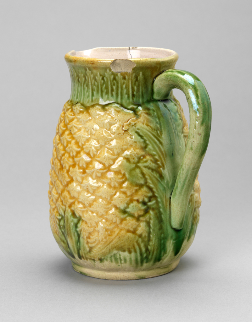 An image of Pineapple jug. Unidentified factory, England. Cream earthenware jug, press moulded in the shape of a pineapple, with a short slightly flaring cylindrical neck, a loop handle and a slim circular foot-rim. The outside is moulded to represent both the fruit and the leaves, the former covered by bright yellow-gold glaze and the latter with a thin green glaze. The interior is tinged pink. Earthenware, press moulded and coated with clear and coloured glazes, height, whole, 14 cm, width, whole, 12 cm, 19th century.