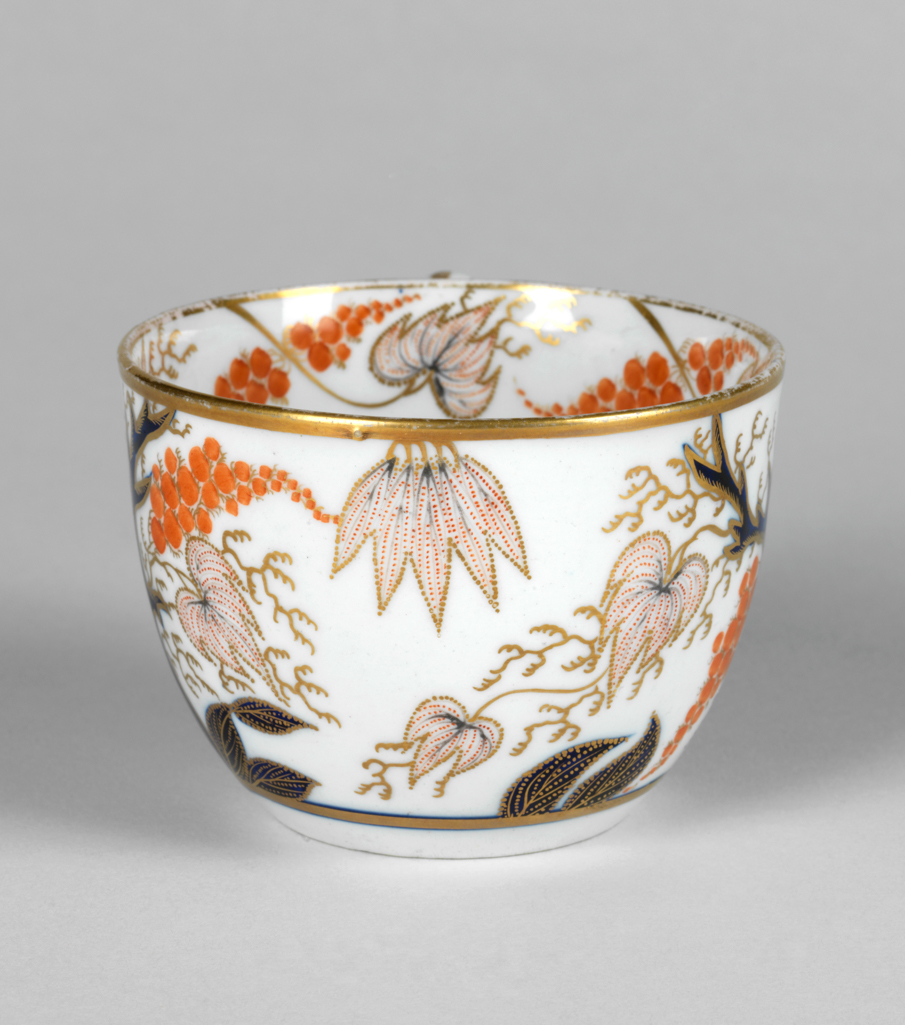An image of Cup and saucer/teacup and saucer. New Hall Porcelain Factory, Staffordshire, New Hall. Oriental style blue trees bearing pinkish-red leaves, bunches of red berries, and gold tendrils. Pattern 446. The circular cup has deep curved sides, a ring handle and a recessed base. The saucer has deep curved sides and stands on a footring. The exterior of the cup is decorated with two blue trees bearing dotted red leaves, bunches of orange berries, and gold tendrils. Inside the rim there are four dotted leaves and four paired bunches of berries, and tendrils, and in the middle, four blue leaves, a bunch of red berries and gold tendrils. There is a gold horizontal band below the design, and round the rim, and a vertical line down the back of the handle. The saucer has a central medallion filled with blue leaves, gold tendrils, and one large red berry surrounded by a blue and gold circle, from which extend two radiating blue trees with leaves and bunches of berries matching those on the cup. A gold band encircles the rim. Hard-paste porcelain painted in blue under presumed lead-glaze, and painted overglaze in orange-red enamel and gold, height, cup, 5.8 cm, width, cup, 10.5 cm, diameter, cup, 8.2 cm, height, saucer, 3 cm, diameter, saucer, 13.8 cm, circa 1785-1810.