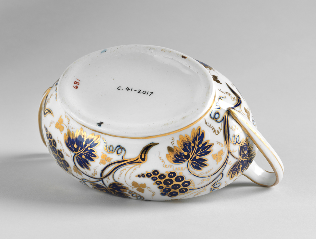 An image of Sugar basin and cover/sugar bowl and cover. New Hall Porcelain Factory, Staffordshire, New Hall. Painted underglaze in mazarine blue with vine leaves, bunches of grapes and tendrils, and gilded. Pattern no. ‘631’ painted in red. Oval with curved sides, and two strap handles which rise upwards before descending to join the lower part of the sides. Inside the top there is a ledge to support the cover. The oval cover rises steeply to the middle where there is a hole, covered by the oval conical knob. The sides of the bowl are decorated with pattern 631 comprising sprays of vine leaves, tendrils and bunches of grapes, and there are gold bands round the lower edge, the top, and the sides of the handles, which also have a longitudinal gold stripe. The cover is decorated en suite but with smaller sprays. It has a wide gold band round the edge and gold knob. Hard-paste porcelain moulded and painted under presumed lead-glaze in mazarine blue and gilded. Transfer-printed underglaze in cobalt blue, height, whole, 12.1 cm, length, whole, 18.8 cm, width, whole, 10.6 cm, circa 1800-1812.