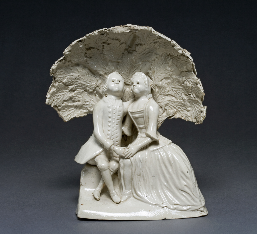 An image of Figure group. Lady and Gentleman in an Arbour. Staffordshire. A lady (on the viewer's right) and a gentleman are seated with their arms around each other under an arbour formed of separately moulded vine leaves. The gentleman has raised her skirt to reveal her right lower leg, and the lady places her hand on his as if to prevent further advances - or perhaps to encourage them? White salt-glazed off-white stoneware, moulded in parts, assembled, and salt-glazed, with brown slip eyes on the figures, height 17.8 cm, circa 1755.