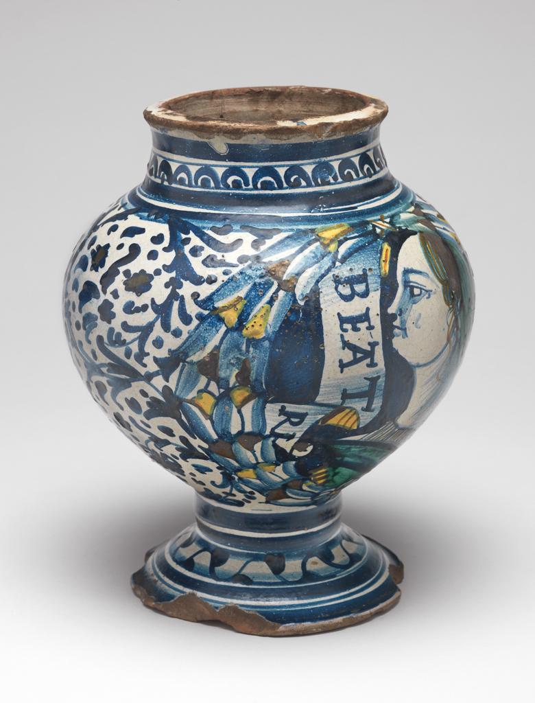 An image of Maiolica Vase. 'Master of the frowning profiles', possibly, Campania, Naples or Vietri. On the front, within a wreath of leaves and berries, is a bust of a dour young woman in profile to left facing a scroll inscribed `BEATRI' (Beatrice). The rest of the body is decorated with stylised scrolling foliage and dots. On the neck, there are semi-circles enclosing spots between horizontal bands; on the stem, two horizontal bands, and on the foot, a wavy line with spots at intervals, and concentric bands of different widths. Pinkish-buff earthenware, tin-glazed on the interior and exterior; base unglazed. Painted mainly in dark blue, with a little green, yellow, and brownish-orange. Height, whole, 21.2 cm, diameter, rim, 10.2 cm, diameter, foot, 12.0cm, diameter, wdiest part, 17.4 cm, circa 1500-1550. Renaissance. Notes: This is one of several bulbous vases and albarelli decorated with frowning profile busts, whose painter was named `Maestro dei profili corruciati' (Master of the frowning profiles) by Donatone (1993, pp. 82-3).