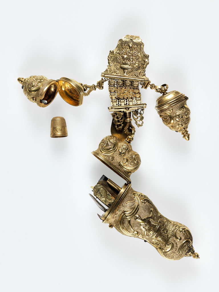 An image of Objets de vertu. Chatelaine with etui and two thimble cases. Gilt metal chatelaine, embossed and chased with rococo decoration. The chatelaine comprises three hinged sections; the uppermost with a long spatulate hook; the central one comprising 13 small linked plaques; the lowest with openwork scrolls either side of a swivel loop. Suspended from the top section are two thimble cases (A & B) with hinged lids. One case contains a thimble (C). Suspended from the lowest section is a shaped triangular etui (D) oval in section, with hinged lid and sprung button clasp. The fitted interior contains a pair of steel scissors (E), a folding knife (F), a silver spoon (G), an earpick/pair of tweezers (H), a pencil case (I) and a pair of ivory memorandum leaves hinged with a gilt button (J). (There are cavities for two more tools, which are missing.) The cast chatelaine is chased with a basket of flowers, the small plaques are chased with a lion and crowns; the embossed etui is chased with two panel containing female figures in contemporary dress within borders of masks, scrolls and rocaille decoration; the thimble cases are similarly decorated. Gilt metal, cast, embossed and chased, length, whole, 20.0 cm, height, etui, 2.5 cm, length, etui, 11.0 cm, width, etui, 3.8 cm, height, thimblecase, 2.3 cm, length, thimblecase, 4.5 cm, width, thimblecase, 2.5 cm, circa 1760. Rococo.