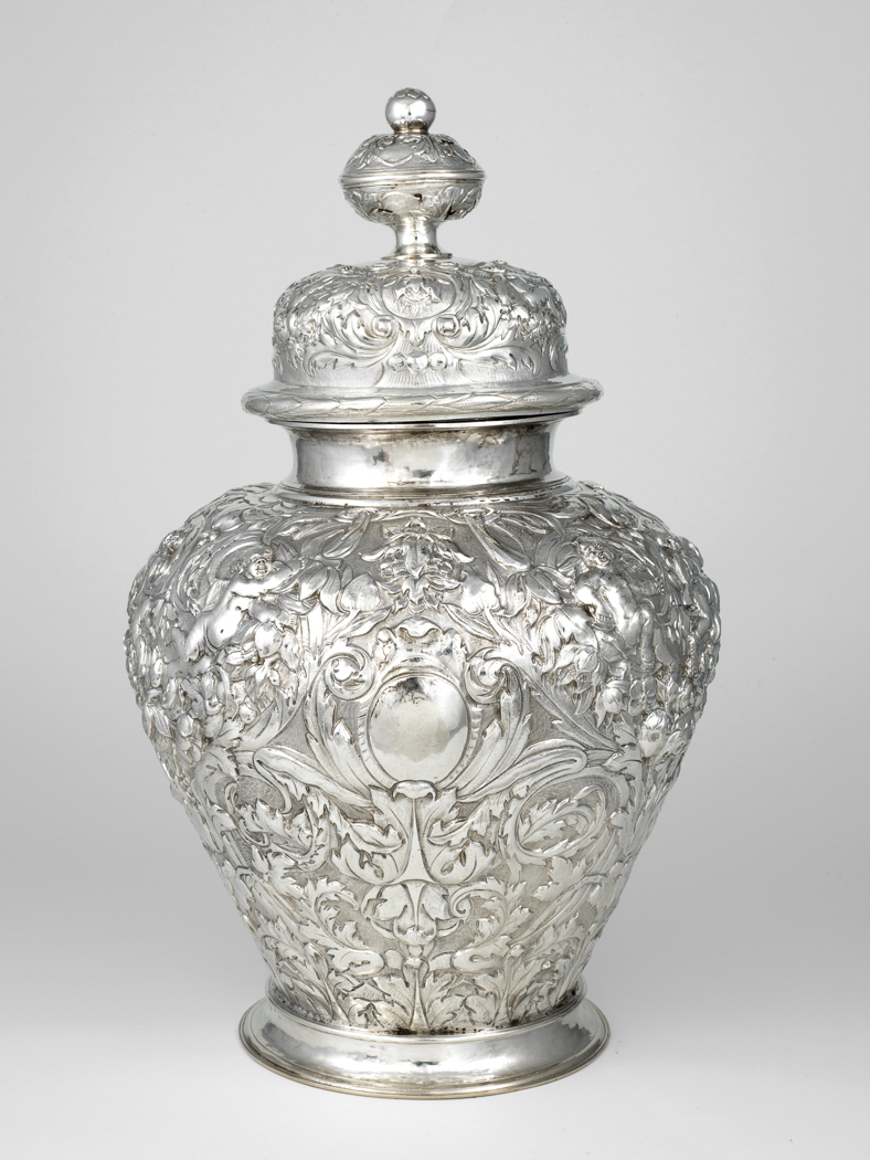 An image of Ginger jar and cover. Bodenendeich, Jacob (Luneburg, 1633/4- London 1681). Putti amongst scrolling foliage against a matted ground; plain neck and foot; replacement knob. Of robust baluster form with incurved neck and foot, domed cover with projecting rim, and a finial comprising a depressed globular knop made in two halves, surmounted by a smaller ball. On three sides of the body there are plain oval cartouches flanked by scrolling foliage. The rest of the surface is decorated in relief with putti amongst scrolling acanthus foliage and flowers against a matted ground. The incurved neck and foot are plain. The projecting edge of the cover is decorated with bound laurel, and the dome with three circular cartouches containing a stylized bud, two of which are flanked by putti with swags below them, and by acanthus foliage which extends round the sides, against a matted ground. In the centre there is a plain silver band from which the finial rises. The depressed globular knop is decorated with upward and downward pointing acanthus foliage, radiating from the bottom and top respectively, and the ball above has a stylized flower on its top. Silver, embossed, chased, and matted, height (whole) 46 cm, diameter (whole) 28.5 cm, weight 2900g, circa 1673-1674. Baroque.