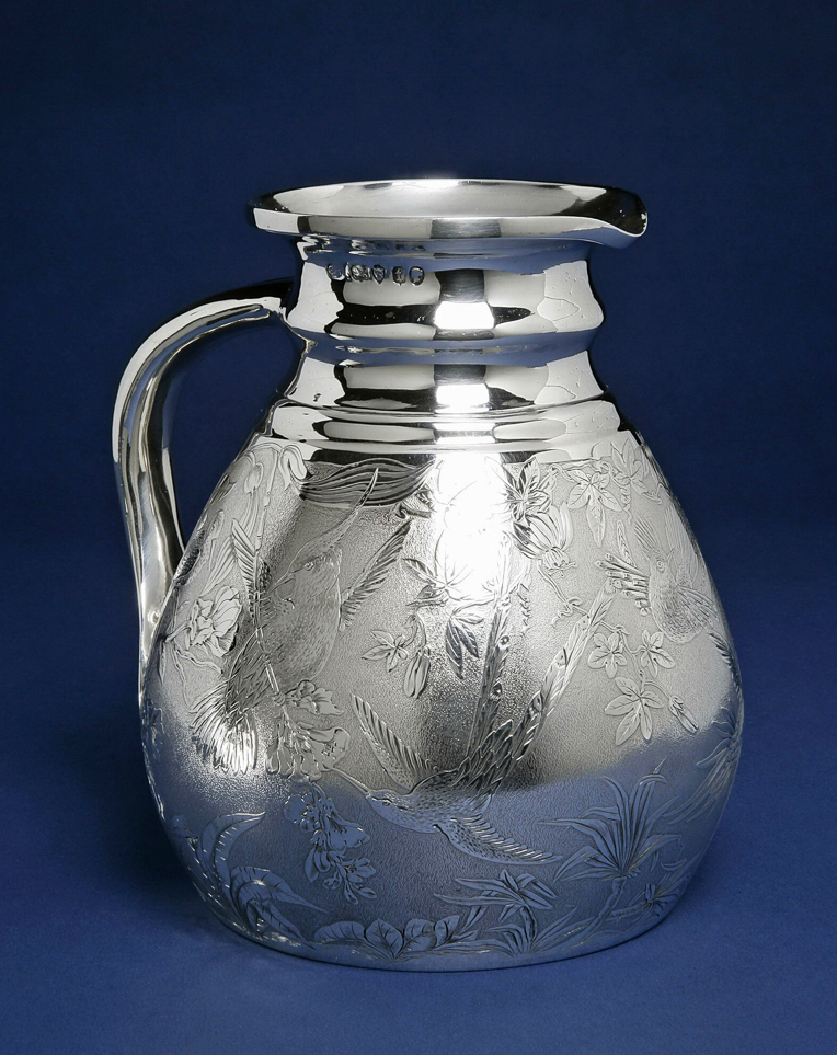 An image of Cambridge ale jug/water jug. Smith, Stephen (British). In the shape of a Cambridge ale jug, the body with sloping cylindrical sides and slightly tucked-in base and flat bottom. The narrow neck with plain moulded rings below a slightly everted rim. The broad handle is concave down the centre and joins the body near the base with a flat spatulate terminal. The sides of the body are matted and engraved with exotic birds and tropical flowers and foliage (including humming birds and passion flowers). The hollow handle was seamed down its length. The body was raised or spun, then the base neck and handle were soldered on. The body was engraved, and the interior gilt. Height, to the rim, 18.2 cm, width, overall, 17 cm, weight, whole, 845 g, 1874-1875. Aesthetic Movement. Victorian. England, London.