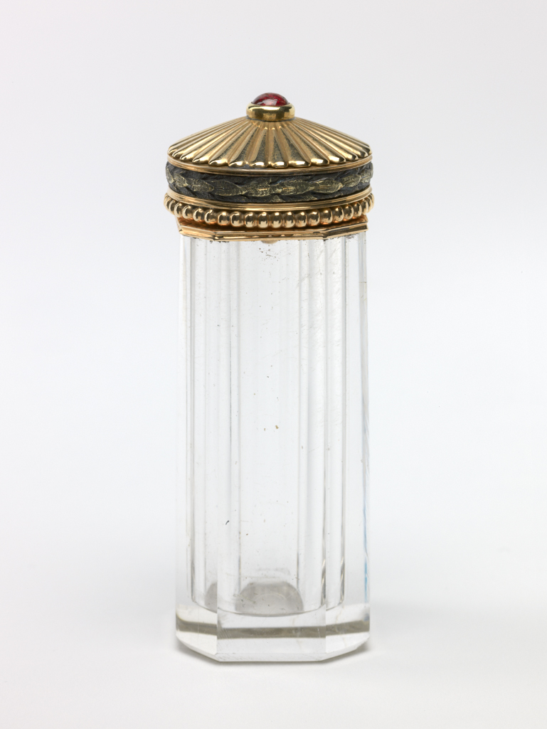 An image of Objets de vertu. Scent bottle/scent flask. Russian. The rock crystal body is of octagonal section with straight sides externally, and cylindrical internally; it has a gold rim mount and hinged lid. The lid has a border of beads round the rim, above which is a chased border of overlapping leaves enamelled in dark translucent green. The top of the lid is slightly domed and is embossed with flutes centering on a cabuchon garnet. Inside is a flat gold stopper (A) with compressed ball-shaped knop and cork base. With fitted case (B). Rock crystal perfume container, gold, enamel and a garnet, height, whole, 2.1, cm, length, whole, 6.0, cm, width, whole, 2.1, cm, circa 1900 to circa 1910.