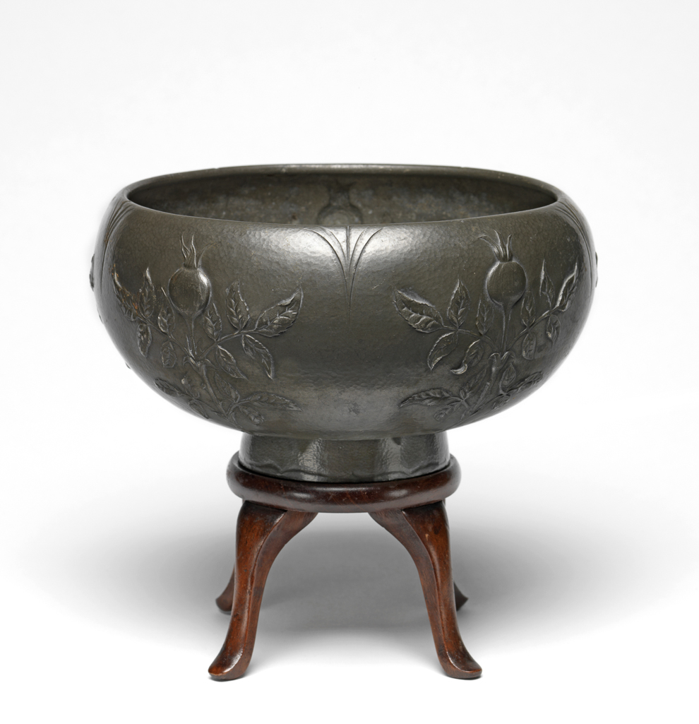 An image of Bowl. Marks, Gilbert Leigh (British, 1861-1905). Circular with deep curved sides, standing on a footring. The sides are decorated with five sprays of leaves, each bearing a rose hip, alternating with five points extending downards from the rim. The edge of the foot is decorated with a cusped line. Circular wood stand with four legs (A). Pewter with lightly hammered surface and embossed decoration, height, bowl, 12.3 cm, diameter, bowl, 22.9 cm, 1901. Arts and Crafts.