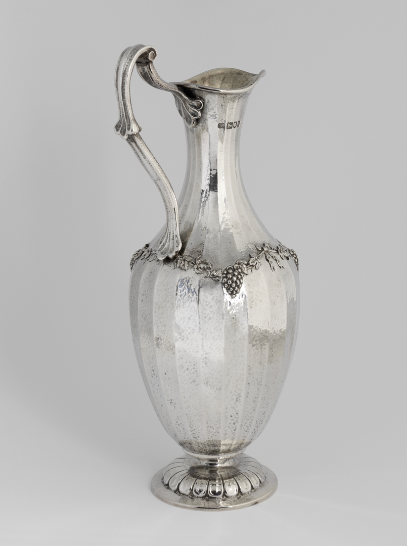 An image of Claret jug. Marks, Gilbert Leigh, silversmith (1861-1905). Facetted, lightly hammered sides decorated with a swag of vine leaves, grapes and ribbons. The tall slender jug has an ovoid body, which is facetted and spot hammered, and is decorated round its shoulders with a swag of vine leaves, grapes on ribbons. It stands on a slightly domed circular food, which is embossed with a border of stylised leaves. The cast harp-shaped handle has stylised shell terminals. Silver, facetted and spot-hammered, with embossed and chased decoration, and cast handle; the interior gilt, height, to top of handle, 30.6 cm, width, overall, 15.4 cm, weight, whole, 860 g, 1898-1899. Arts and Crafts.