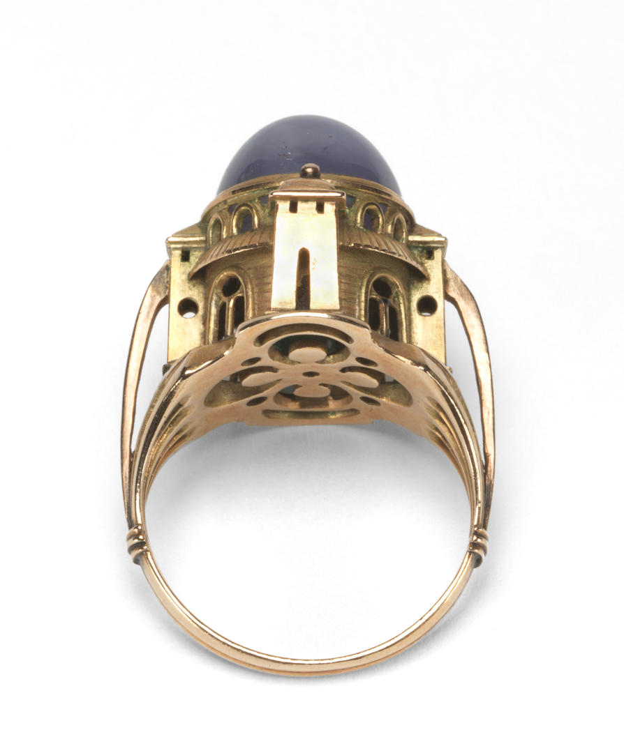 An image of Jewellery/Ring. The Sabbatai Ring. Ricketts, Charles de Sousy, designer (British, 1866-1931). Gold ring with a mosque-shaped bezel, the dome formed by a cabochon star sapphire, the hollow interior containing a loose emerald. The shoulders of the ring are each pierced by four long tapering holes, and the bezel is in the form of a mosque with tower-like buttresses, four pierced doorways, each with a sloping roof and two arched windows above. The doors were originally smeared with ambergris. Gold, set with a cabochon star sapphire, and containing a loose emerald, 1904. Edwardian. Notes: Ricketts promised to make a ring for Miss Bradley in December 1903. The form was inspired by the story of Sabbatai Sebi (1626-76), a Jewish mystic, who having proclaimed himself Messiah in Jerusalem, was later converted to Islam. It represents the mosque of Omar, and was intended to appeal to the five senses. See Documentation, Scarisbrick, 1982.