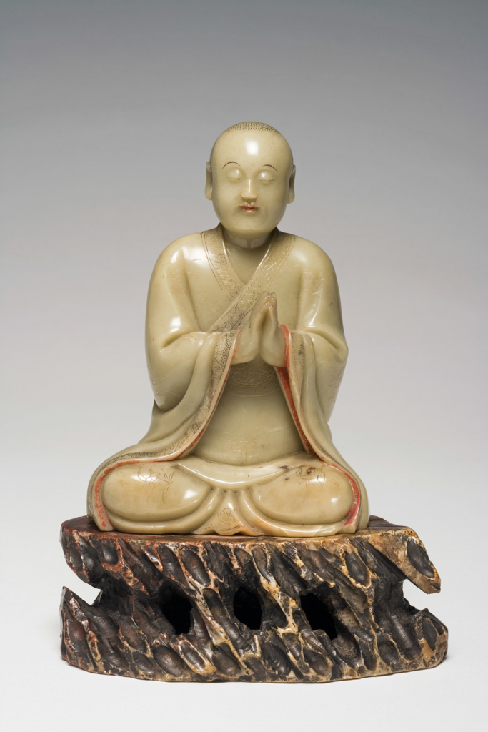 An image of Carving. Soapstone, carved as a seated luohan with his eyes closed and his hands together in a respectful gesture. The collar, sleeves and hem of his robes incised with a design of scrolling lotus on a formalized wave ground. Soapstone, height, including base, 17.5 cm, 1700-1800, Chinese.