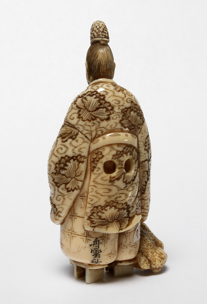 An image of Netsuke. Ono No Tofu in his ceremonial garments wearing Geta (wooden clogs) standing and holding his umbrella in his right hand and a folding fan in his left hand. A large frog is sitting next to his right foot. Ivory, carved, the eyes of frog are inlaid with horn, 1870-1900. Japanese.