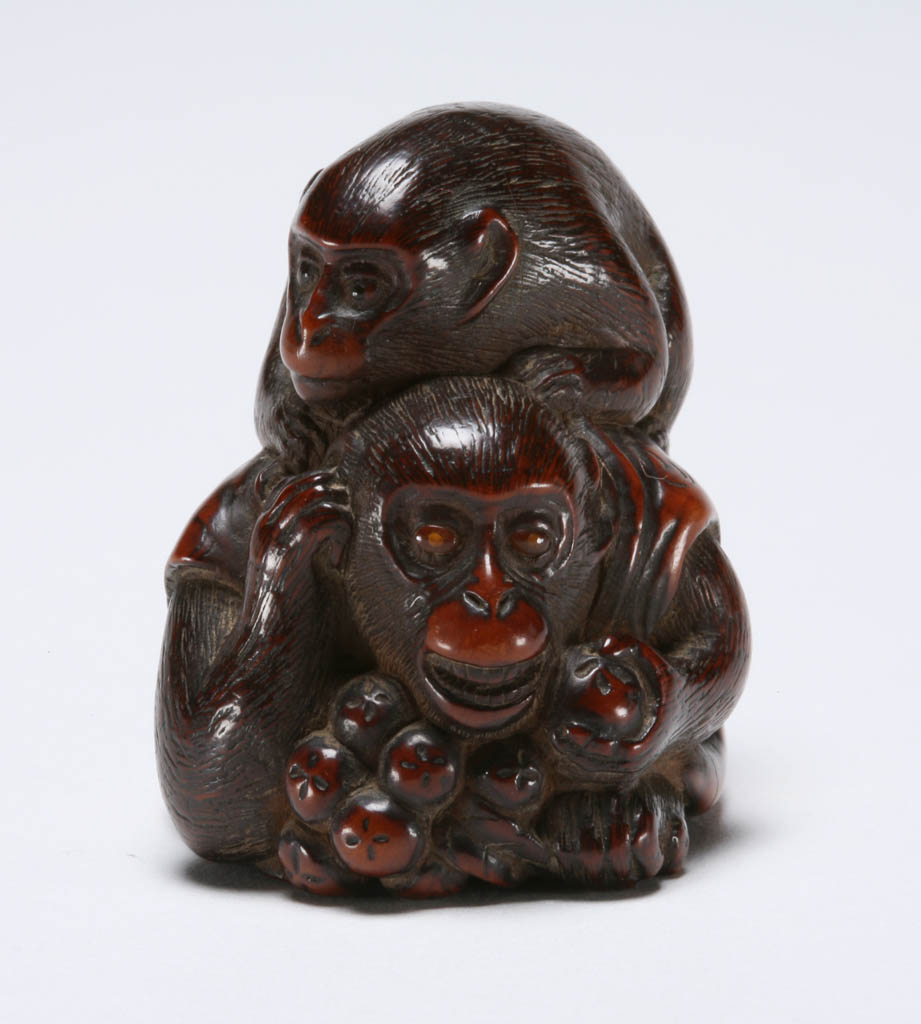 An image of Netsuke. A monkey wearing a vest crouching down and enjoying a large branch of loquats, a younger monkey on its back trying to distract the older monkey for some loquats. Boxwood, carved, eye single and double inlaid in horn, 1800-1850. Edo Period.