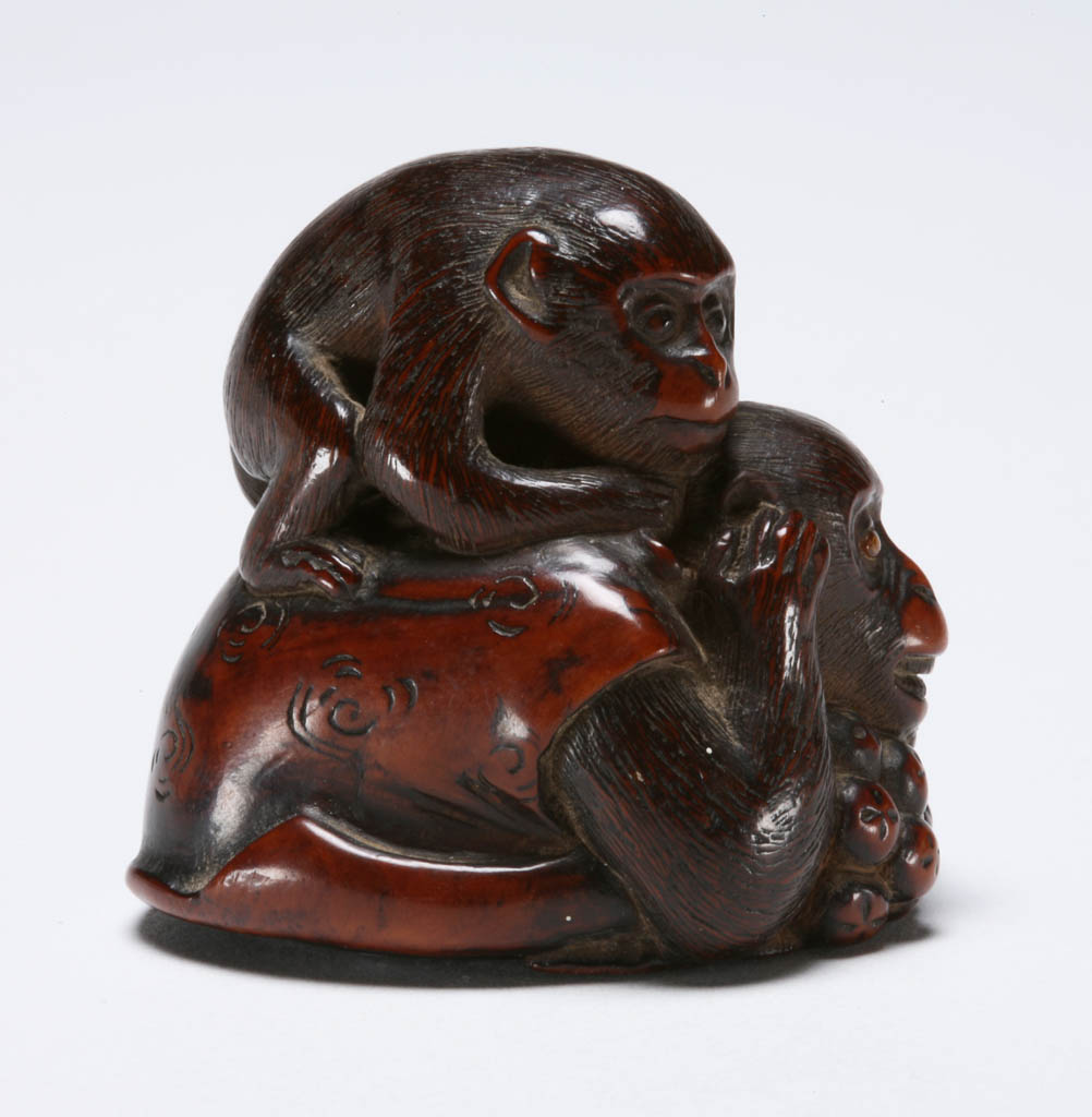 An image of Netsuke. A monkey wearing a vest crouching down and enjoying a large branch of loquats, a younger monkey on its back trying to distract the older monkey for some loquats. Boxwood, carved, eye single and double inlaid in horn, 1800-1850. Edo Period.