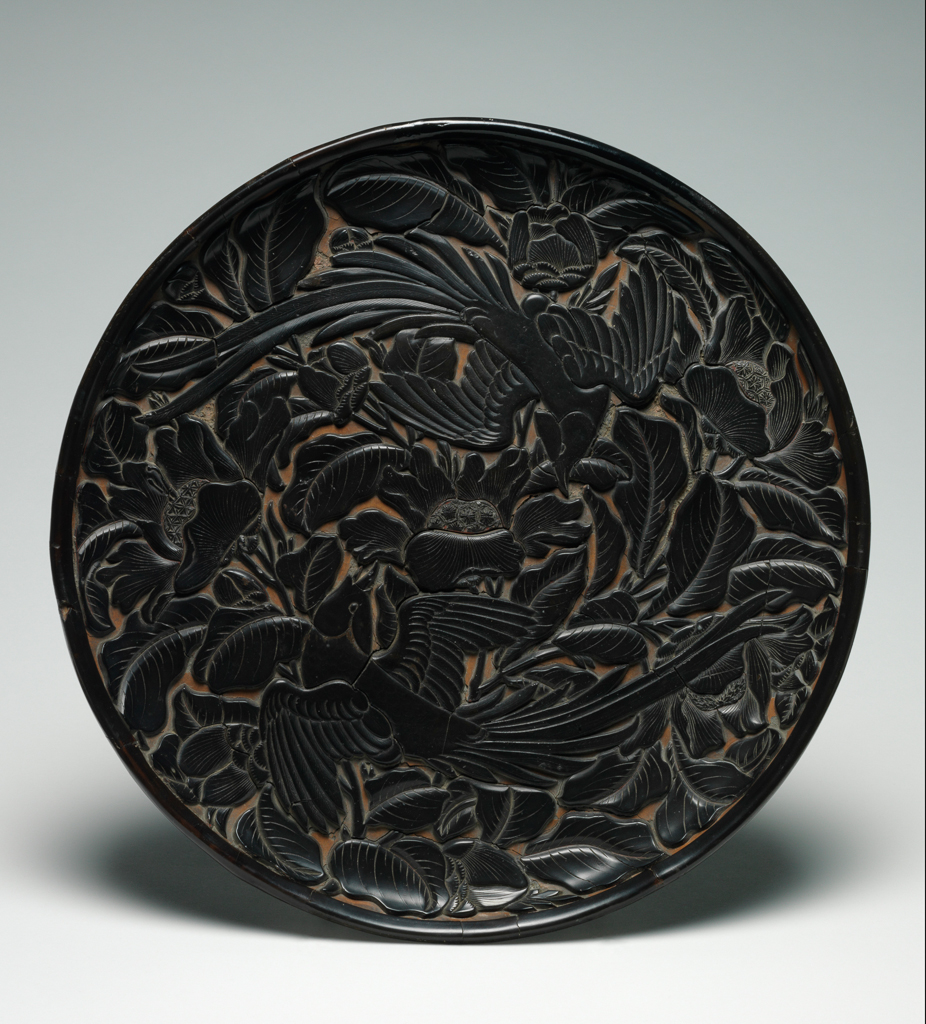 An image of Dish. Circular dish, with deep carving through black lacquer to a buff ground. Depicting two long tailed birds amid foliage and flowers. Carved, diameter 32 cm, 14th century. Yuan Dynasty (1279-1368). Chinese.