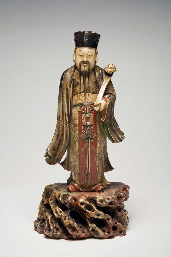An image of Soapstone carved as a Star God of Rank and Affluence in the form of a long-bearded official, dressed in an elaborate hat and long flowing robes. He is holding a ruyi sceptre in the left hand. The robes are intricately carved with elaborate floral designs. Soapstone, carved, height 38, cm, width, 19.2 cm, 1700-1900. Chinese.