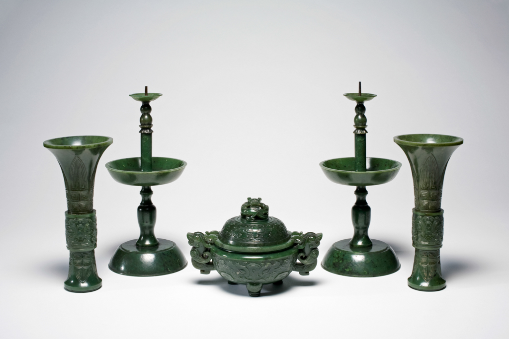 An image of Altar set of five pieces. Dark green with black spots; a Buddhist altar set of five pieces; two pricket candlesticks, two vases, and an incense burner. The two vases and incense burner are carved with ancient jade designs. The set are mounted on an elaborately carved wooden stand. Jasper, 1700-1800. Chinese.