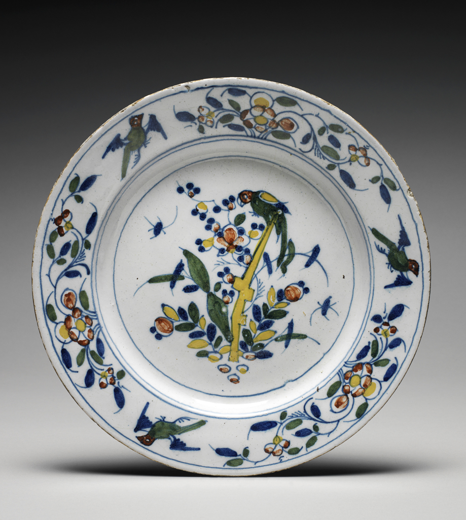 An image of English Delftware. Plate. Lambeth Pottery, probably. The middle is decorated with flowering plants, a parrot perching in a tree, and flying insects; on there are three flying birds alternating with three stylized floral sprays. Earthenware, moulded, tin-glazed, painted in high-temperature colour, diameter 21.9 cm, circa 1750-1775.