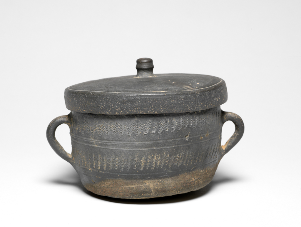 An image of Stoneware bowl. Unknown maker. High fired, well-ringing bucket-shaped bowl with two loop handles, bevelled rim, and shallow, well-fitting knobbed lid, the bottom untidily convex, of dirty-grey body with black wash-like tint ash glaze, with two rows of narrow-combed decoration. Inside the lid a long character inscription. Before (?) AD 700. Korean?