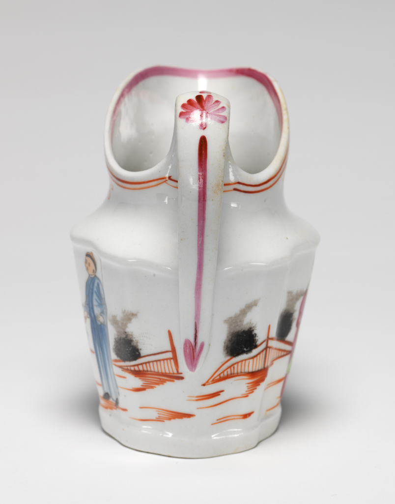 An image of Milk Jug. New Hall Porcelain Factory, Staffordshire. Painted with a Chinese scene known as The Boy with a Butterfly. Of silver-shape with a loop handle. The exterior is decorated with a continuous Chinese scene: two fuzzy black bushes behind a red fence; a woman wearing green and puce clothing, standing holding a sceptre; a black bush and a rock beside a flowering tree; a boy dressed in green and dark puce who is holding up his arms towards a butterfly; two black bushes flanking a flowering tree; a standing man dressed in blue; and a black bush beside a red fence. The ground is indicated by horizontal red strokes. Round the outside of the rim there are two narrow red lines under which are four inverted hearts with a circle below each. On the inside edge of the rim there is a puce band, and down the back of the handle, graduated dots, a fan-shaped flower, and a long vertical stroke terminating in an arrowhead. Pattern 421. Hybrid hard-paste porcelain, lead-glazed, moulded, and painted in enamels, height, whole, 10.7 cm, width, whole, 11.6 cm, circa 1800-1805. Chinese Style.