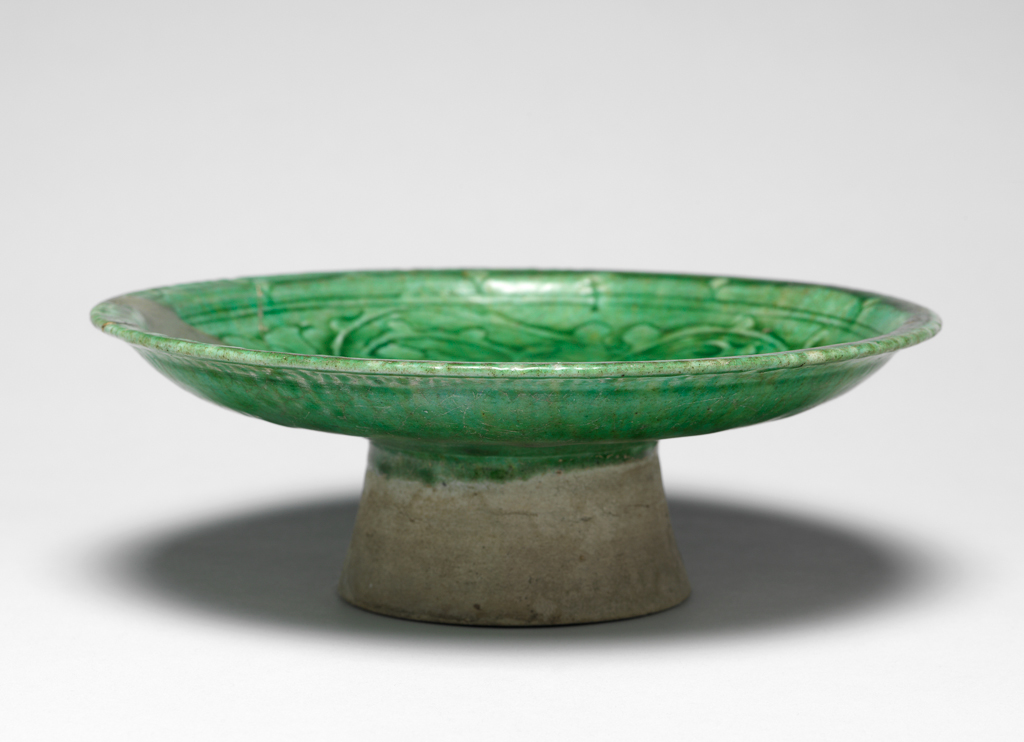 An image of Islamic Pottery. Dish. Monochrome glazed ware. Tazza or dish comprising circular shallow dish with everted rim, sitting on a high foot ring with slightly flaring profile. Carved decoration fills the interior and is covered with a crazed, translucent green glaze. The pattern on the rim incorporates repeating diamond pattern alternating with scalloped line which is separated from the principal decorative theme by two parallel concentric lines. A carved repeating and curving arabesque pattern forms a circular frieze on the body. A quatrefoil rosette fills the base. Buff fritware, wheel thrown, with carved decoration and a translucent green glaze, height, whole, 6.9 cm, width, whole, 20.3 cm, diameter, rim, 20.3 cm, diameter, base, 8.7 cm, weight, whole, 409 g, circa 1100-1300. Ayyubid Dynasty. Syrian.