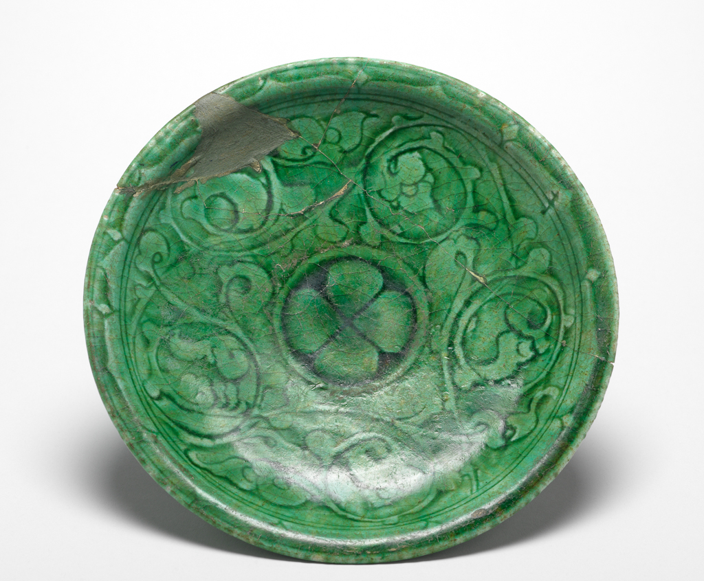 An image of Islamic Pottery. Dish. Monochrome glazed ware. Tazza or dish comprising circular shallow dish with everted rim, sitting on a high foot ring with slightly flaring profile. Carved decoration fills the interior and is covered with a crazed, translucent green glaze. The pattern on the rim incorporates repeating diamond pattern alternating with scalloped line which is separated from the principal decorative theme by two parallel concentric lines. A carved repeating and curving arabesque pattern forms a circular frieze on the body. A quatrefoil rosette fills the base. Buff fritware, wheel thrown, with carved decoration and a translucent green glaze, height, whole, 6.9 cm, width, whole, 20.3 cm, diameter, rim, 20.3 cm, diameter, base, 8.7 cm, weight, whole, 409 g, circa 1100-1300. Ayyubid Dynasty. Syrian.