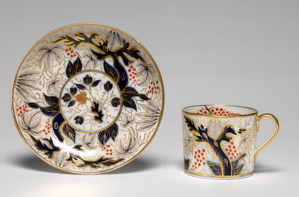 An image of Coffee cup and saucer. New Hall Porcelain Factory, Staffordshire. Cylindrical coffee can with loop handle. Circular saucer with curved sides, standing on a footring. The cup is decorated on the exterior with a blue tree, with foliage dotted in blue and red, and with two bunches of red berries, and gold tendrils, repeated on the other side. Inside there are two gold branches, each with two dotted leaves, and two bunches of red berries. There are gold bands round the rim and base and the back of the handle is gilded. The saucer has a central gold-edged medallion containing a plant, surrounded by the pattern on the exterior of the cup. The edge is encircled by a gold band. Pattern no. 372. Hybrid hard-paste porcelain, painted underglaze in enamels,  decoration in cobalt blue, and gilded, height, cup, 5.9 cm, width, cup, 8.8 cm, diameter, cup, 6.8 cm, height, saucer, 3.1 cm, diameter, saucer, 13.6 cm, circa 1795-1800. Oriental Style.