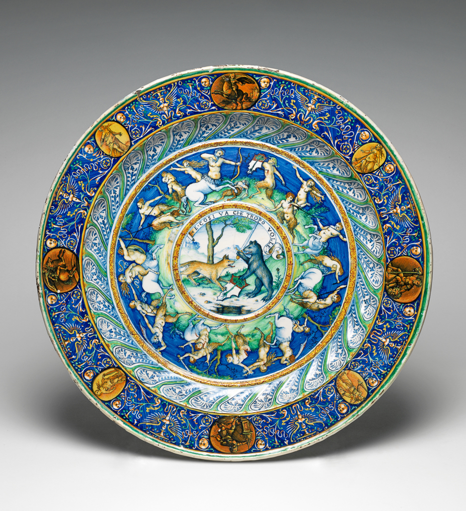 An image of Maiolica Basin. Vulcan Painter. Maiolica basin for ewer, painted in polychrome, with a fox and bear with a shield of arms in a landscape, a battle between centaurs and satyrs, and eight medallions painted with subjects from the Bible and Roman history surrounded by grotesques. Pale buff earthenware, tin-glazed white overall. Height (whole) 5.5 cm, diameter (whole) 46.3 cm. circa 1520 to 1530.