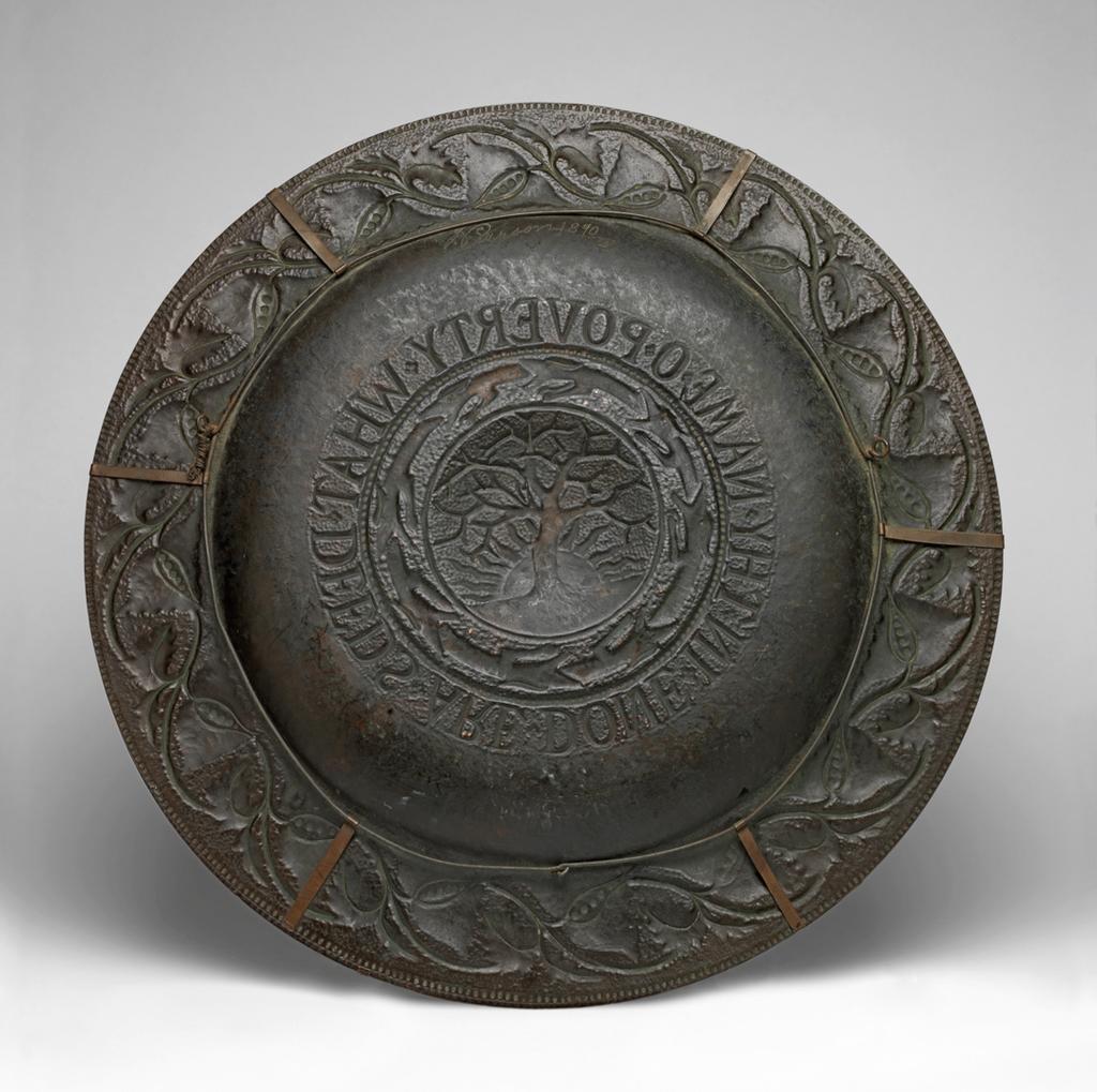 An image of Metalwork. Copper Charger. Pearson, John (British, 1859-1930).  Large, circular, dark brown copper charger, with a wide flat bowl surrounded by a horizontal rim and covered with repoussé decoration. On a raised dome in the centre is a tree, with the sun behind it, surrounded by a circular chain of eight hounds, and around that the inscription ‘WHAT DEEDS ARE DONE IN THE NAME OF POVERTY’. Around the rim is a continuous scrolling design of stylised leaves, some supporting berries. Six point hanging frame fitted around the back. Copper sheet, raised, embossed and chased,  height 3 cm, diameter 51.2 cm, 1890. Arts and Crafts. Lent to the museum by The Keatley Trust.