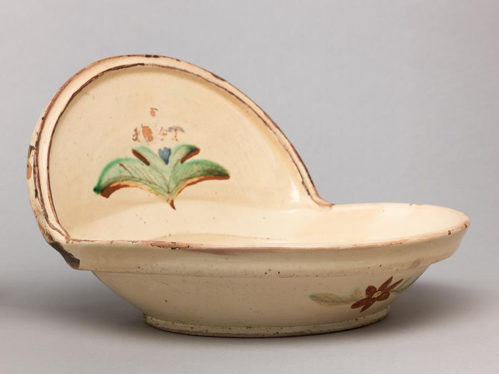 An image of Swiss Slipware. Basin. Lötscher, Andreas (Swiss). Production Place: Switzerland, Canton Graubünden, Sant Antönien. Circular, wth shallow curved sides, and at the back, a vertical arched extension. The front of the exterior is decorated with a red flower flanked by leaves, and there is a plant in the middle of the vertical back, and the date 1841 (the second digit obliterated). Red earthenware, the front only coated with white slip and painted in brownish-red, green, and blue under lead-glaze. On the back some slip has run over from the front, but it is otherwise undecorated. Height 16.5 cm, width, whole, 24.5 cm, dated 1841.