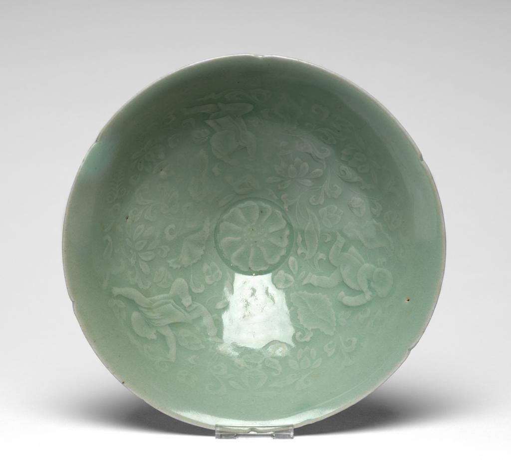 An image of Bowl with boys among lotus design. Unknown pottery, Korea, South Cholla province, Kangjin-gun, Sadang-ri kilns. This conical bowl has six indentations in the rim, suggesting a flower-like shape. The footring is low, the outside is plain, and the inside is finely moulded in shallow relief with a chrysanthemum flower-head in the centre and three boys playing among scrolling lotus round the sides. The pale bluish-green jade-like glaze is evenly applied, with a subtle gloss and no crackle. The base shows three neat quartzite spur-marks. Stoneware, thrown, moulded and celadon-glazed. Height, bowl, 6.7 cm, diameter, rim, 18.9 cm, diameter, foot, 6.2 cm, circa 1100-1150. Koryo Dynasty.