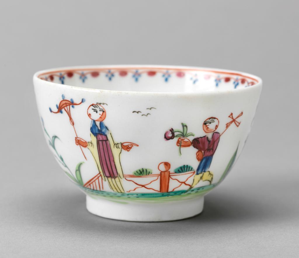 An image of Tea bowl and saucer/cup and saucer. New Hall Porcelain Factory, Staffordshire. Chinoiserie scenes. Pattern no. 20. The circular cup has deep curved sides and stands on a footring. The saucer has deep curved sides and stands on a footring. The cup is decorated on one side with a lady holding a parasol and a boy walking towards her holding a windmill and a flower with a fence behind them. On the right there is a plant in a pot and a small pine bush, and on the left a plant with a blue flower. On the other side there is a mound of grasses and five birds in the sky. Inside the rim is a border comprising a red line, a mauve scalloped line with blue flowers on the points and a dark pink spots on the curves. In the middle there is a small green plant with a purple bud. The saucer is decorated inside with the same figures and flanking motifs on a larger scale, and has a matching border round the rim. Hybrid hard-paste porcelain painted overglaze in blue, two-shades of green, yellow, red, dark pink, pinkish-purple, and black enamels (polychrome). Height, cup, 4.8 cm, diameter, cup, 8.1 cm, height, saucer, 3.4 cm, diameter, saucer, 13 cm, circa 1785-1800. Chinoiserie.