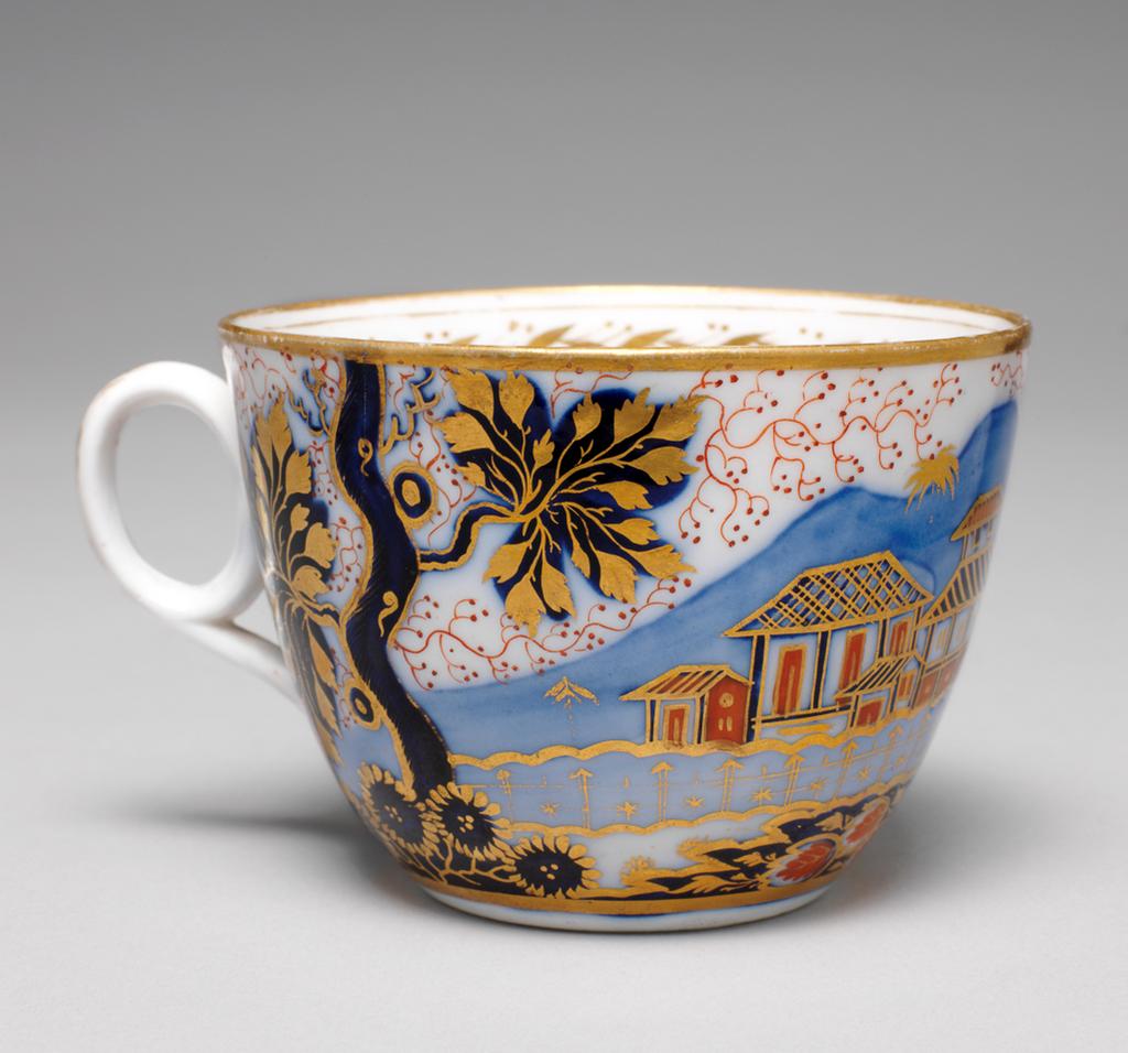 An image of Cup and Saucer/Teacup. New Hall Porcelain Factory, Staffordshire. Bute shape cup with oval ring handle. Circular saucer with curved sides, standing on a footring. The exterior pf the cup is decorated with an Oriental landscape in which are a tree, a group of buildings, a bird perching in a tree branch, and a tiger lying down in front of a rock covered in daisy-like flowers. There is a gold band round the base and rim, inside which is a border of slanting leaves and pairs of berries with a narrow and a wider band above and below. The back of the handle is decorated with five stylized leaves of graduated size. The saucer is decorated with a different version of the design with the tiger lying down on the left below the tree, and with a matching border. Pattern 1214. Bone china, painted underglaze in blue, overglaze in pale apricot and red enamels, and gilded. Circa 1815-1820. Imari style.