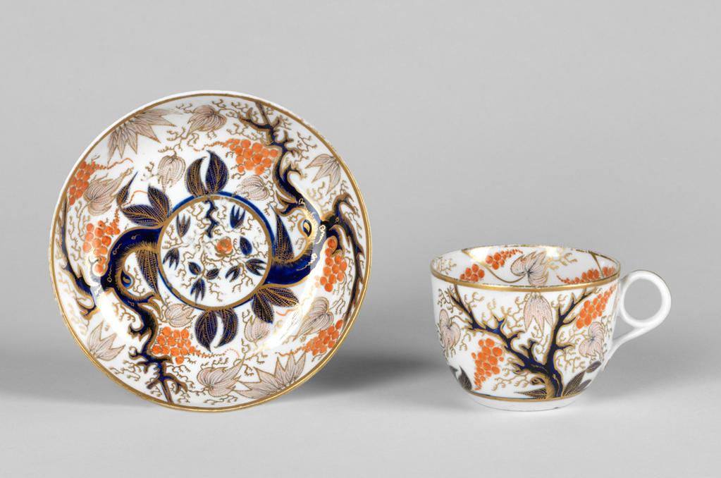 An image of Cup and saucer/teacup and saucer. New Hall Porcelain Factory, Staffordshire, New Hall. Oriental style blue trees bearing pinkish-red leaves, bunches of red berries, and gold tendrils. Pattern 446. The circular cup has deep curved sides, a ring handle and a recessed base. The saucer has deep curved sides and stands on a footring. The exterior of the cup is decorated with two blue trees bearing dotted red leaves, bunches of orange berries, and gold tendrils. Inside the rim there are four dotted leaves and four paired bunches of berries, and tendrils, and in the middle, four blue leaves, a bunch of red berries and gold tendrils. There is a gold horizontal band below the design, and round the rim, and a vertical line down the back of the handle. The saucer has a central medallion filled with blue leaves, gold tendrils, and one large red berry surrounded by a blue and gold circle, from which extend two radiating blue trees with leaves and bunches of berries matching those on the cup. A gold band encircles the rim. Hard-paste porcelain painted in blue under presumed lead-glaze, and painted overglaze in orange-red enamel and gold, height, cup, 5.8 cm, width, cup, 10.5 cm, diameter, cup, 8.2 cm, height, saucer, 3 cm, diameter, saucer, 13.8 cm, circa 1785-1810.
