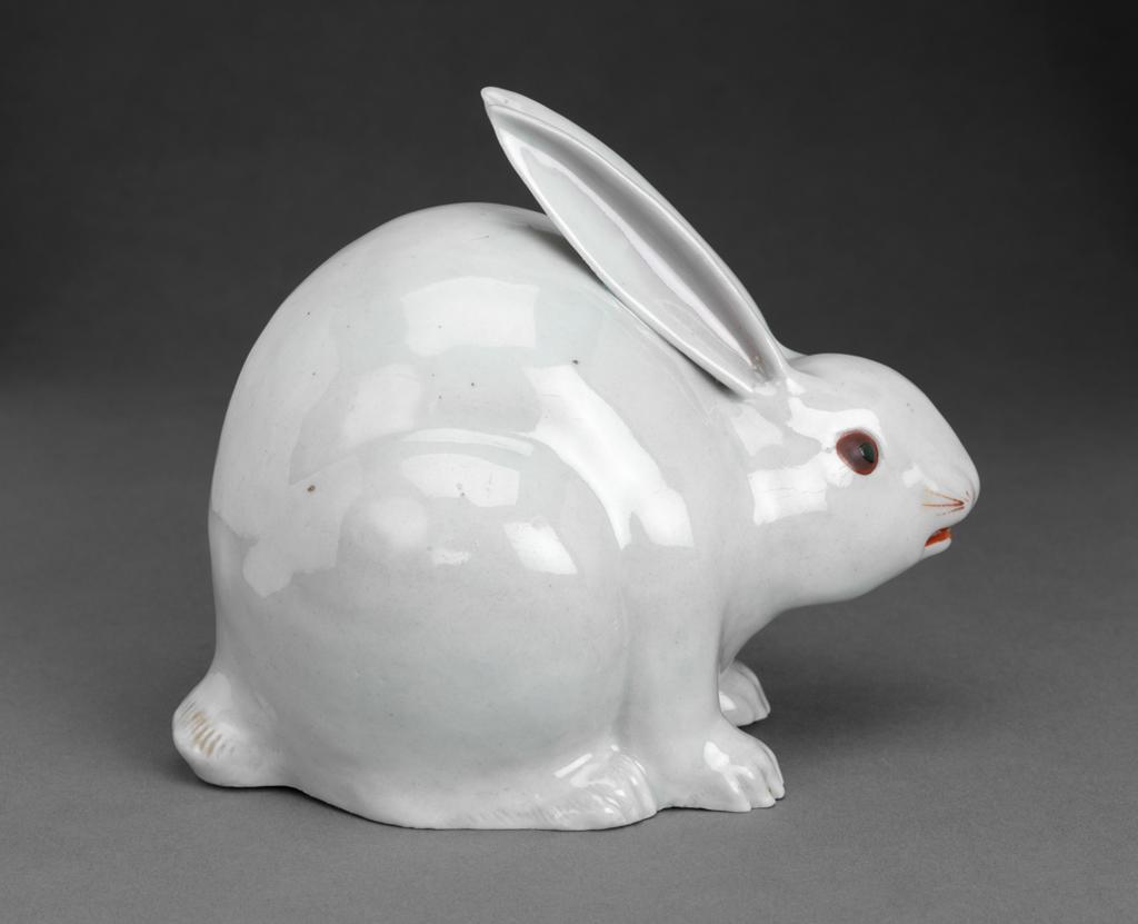 An image of Hare. Hard paste porcelain with greyish glaze and pinkish gold-lustre wash details picked out in red and dark brown and black enamels.  Model of a crouching hare, moulded with hollow interior, the pointed ears, painted with pink-gold lustre on the inside, resting on its arched back, the mouth pierced at either corner and picked out in red enamel; red whiskers; red eyes with dark green pupils; paws and tail with incised details and pinky wash.